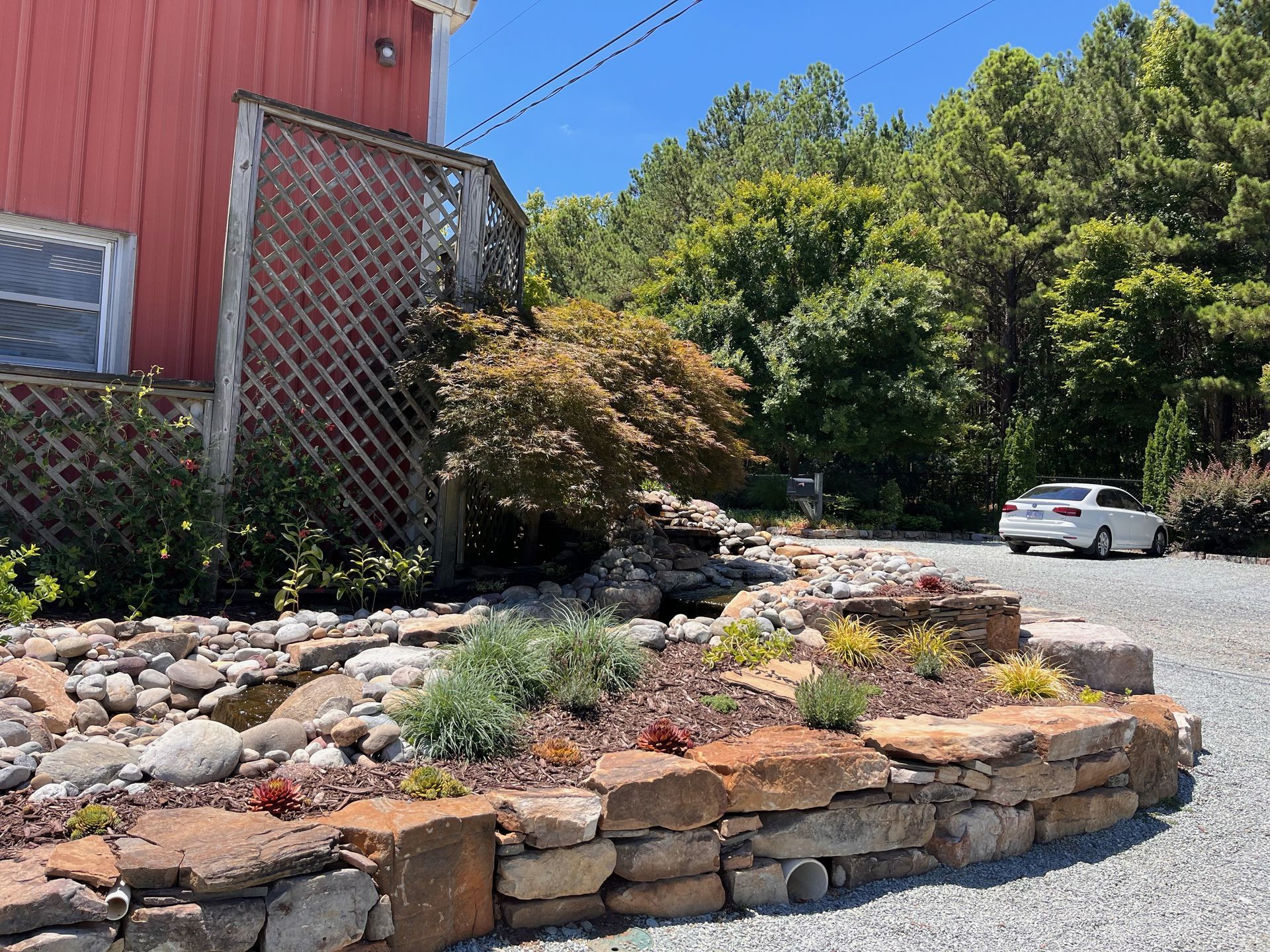 Red barn with stone retaining wall, small pond, and car parked on gravel driveway.
