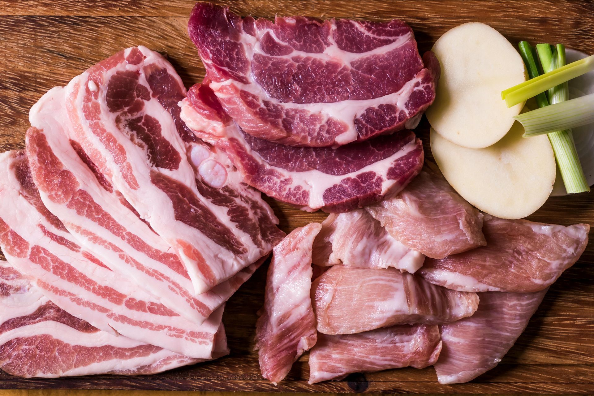 Assorted raw marbled beef cuts arranged on a wooden board with onion slices and green onion stalks