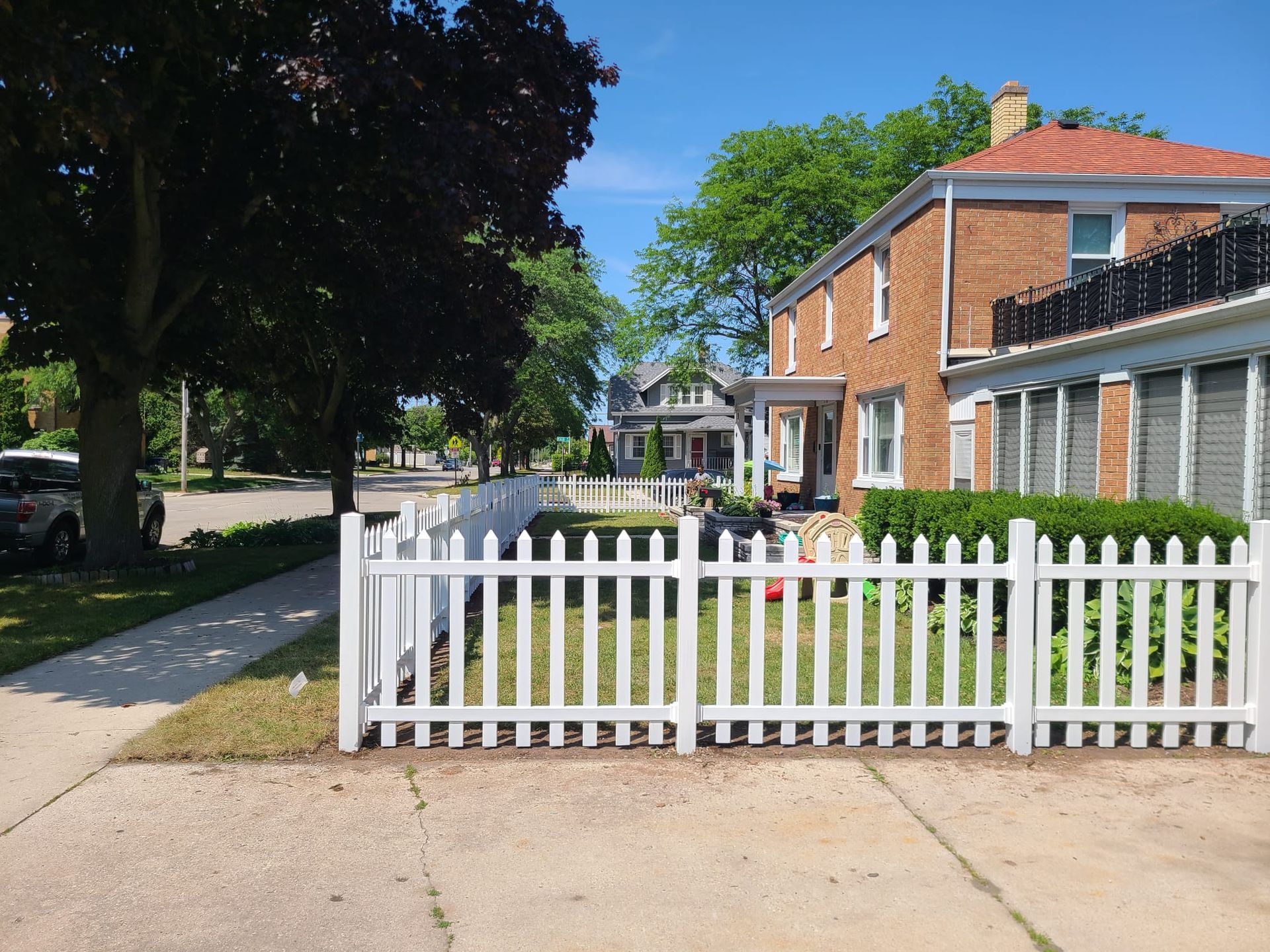A white picket fence runs along a sidewalk beside a brick house and a large tree under a bright blue sky.