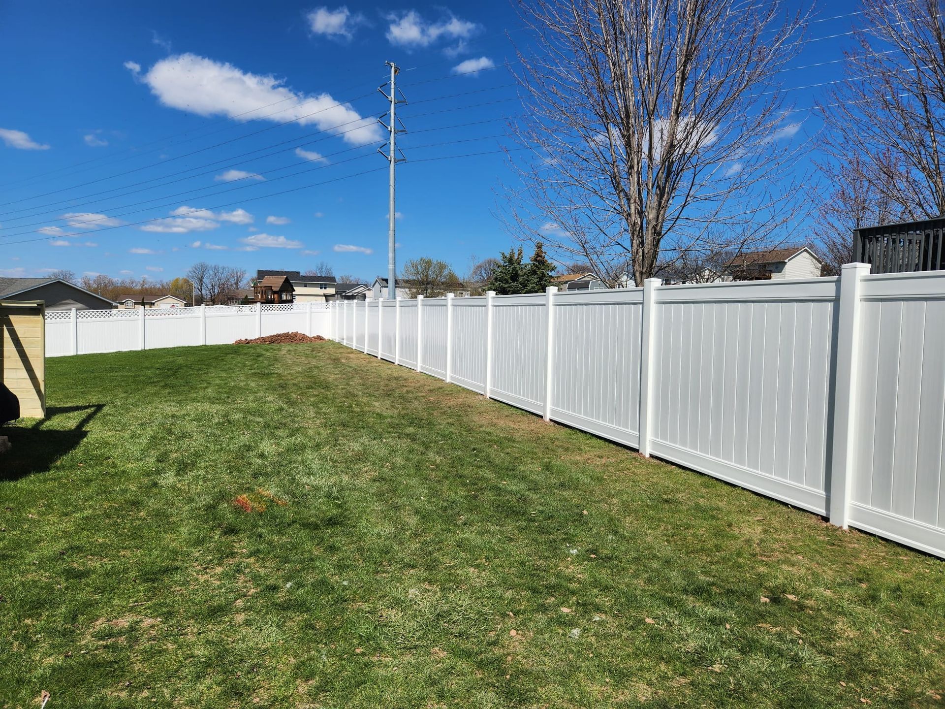 A white vinyl privacy fence lines the edge of a green backyard under a bright blue sky with sparse clouds.