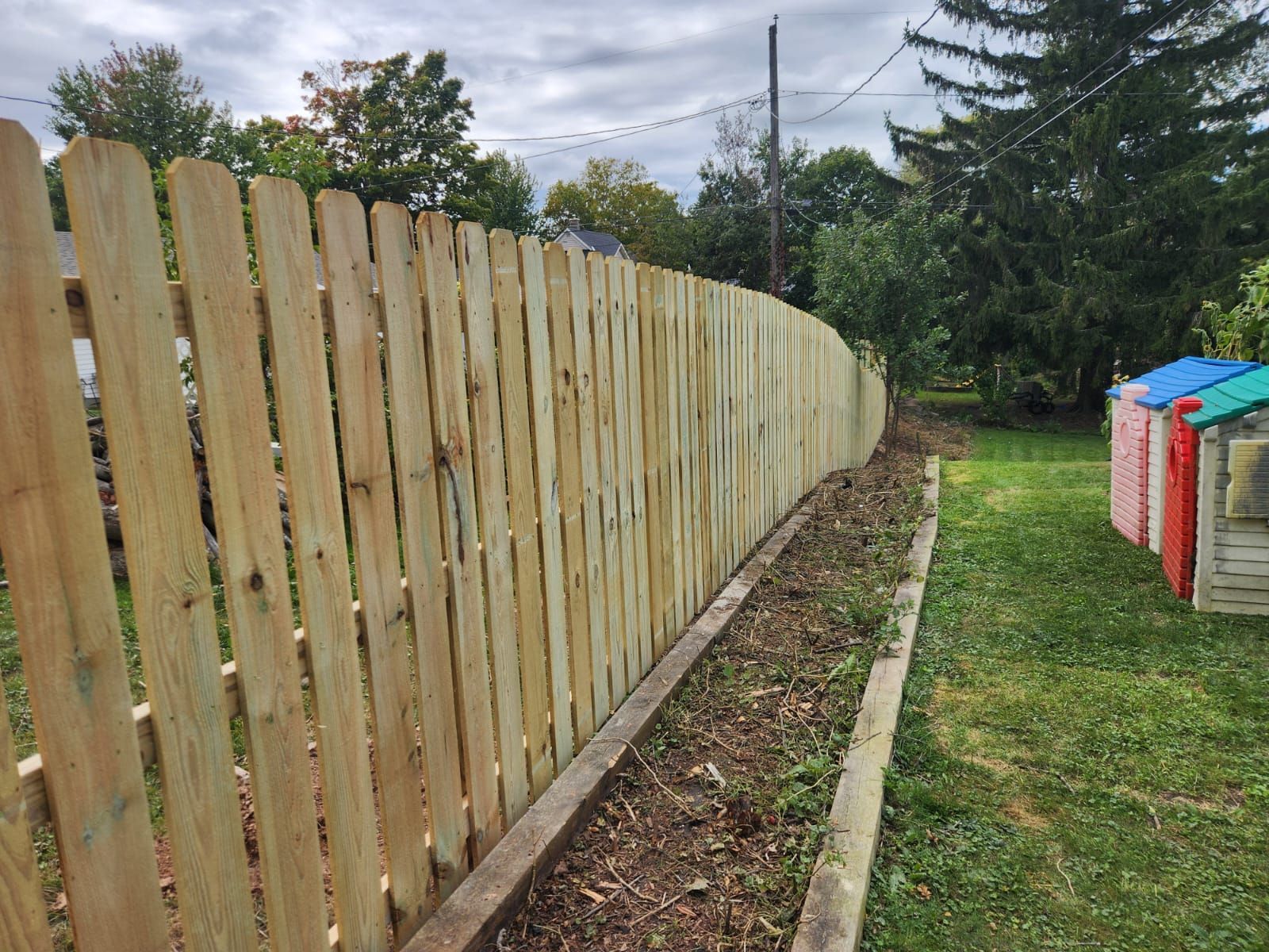 A long, new wooden picket fence stretches across a backyard, next to a garden bed and a row of colorful plastic sheds.