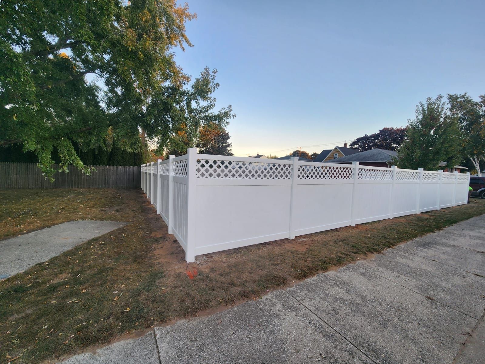 A white vinyl privacy fence with a lattice top sits along a sidewalk next to a grassy area under a clear sky.