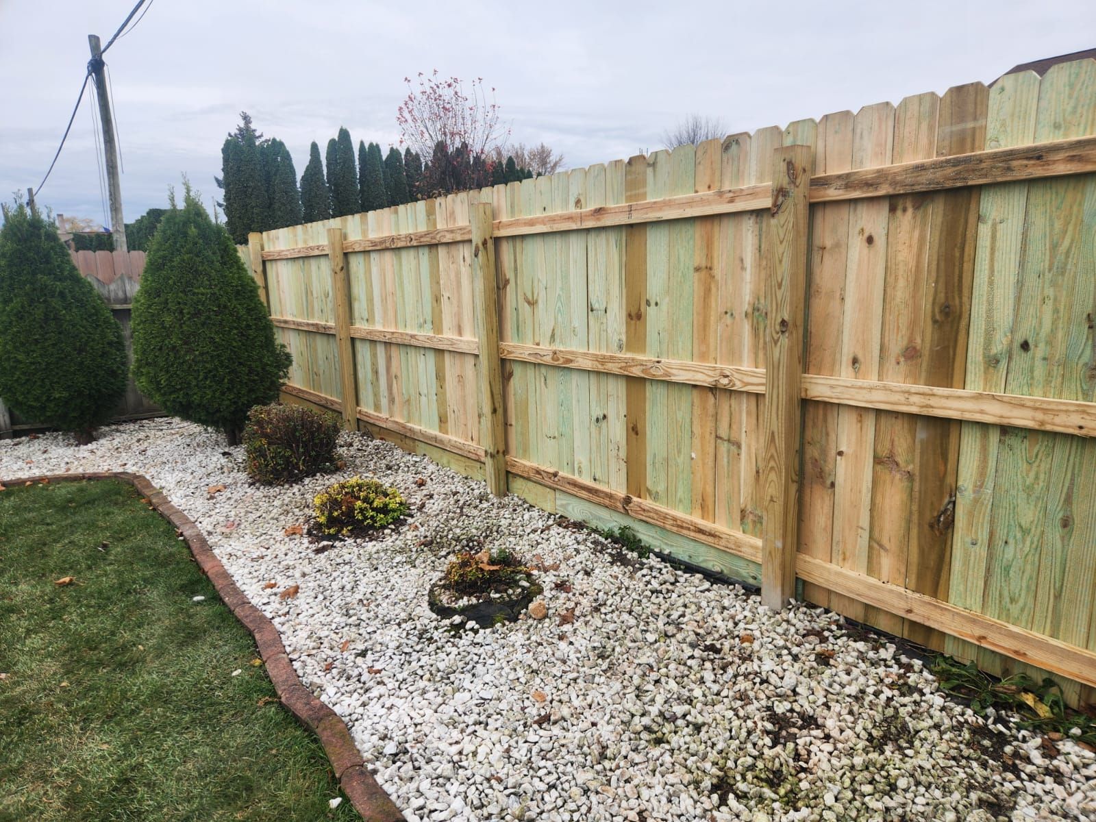 A new wooden fence stands behind a garden bed with white gravel, small shrubs, and a curved brick border.