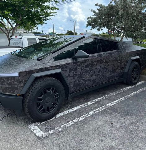 A black car is parked in a parking lot with trees in the background.