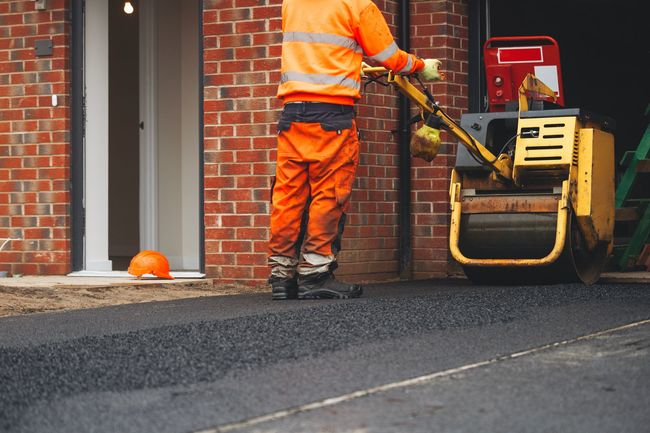 Worker in orange safety gear paving a driveway beside a brick house with a road roller nearby Worker in orange safety gear paving a driveway beside a brick house with a road roller nearby