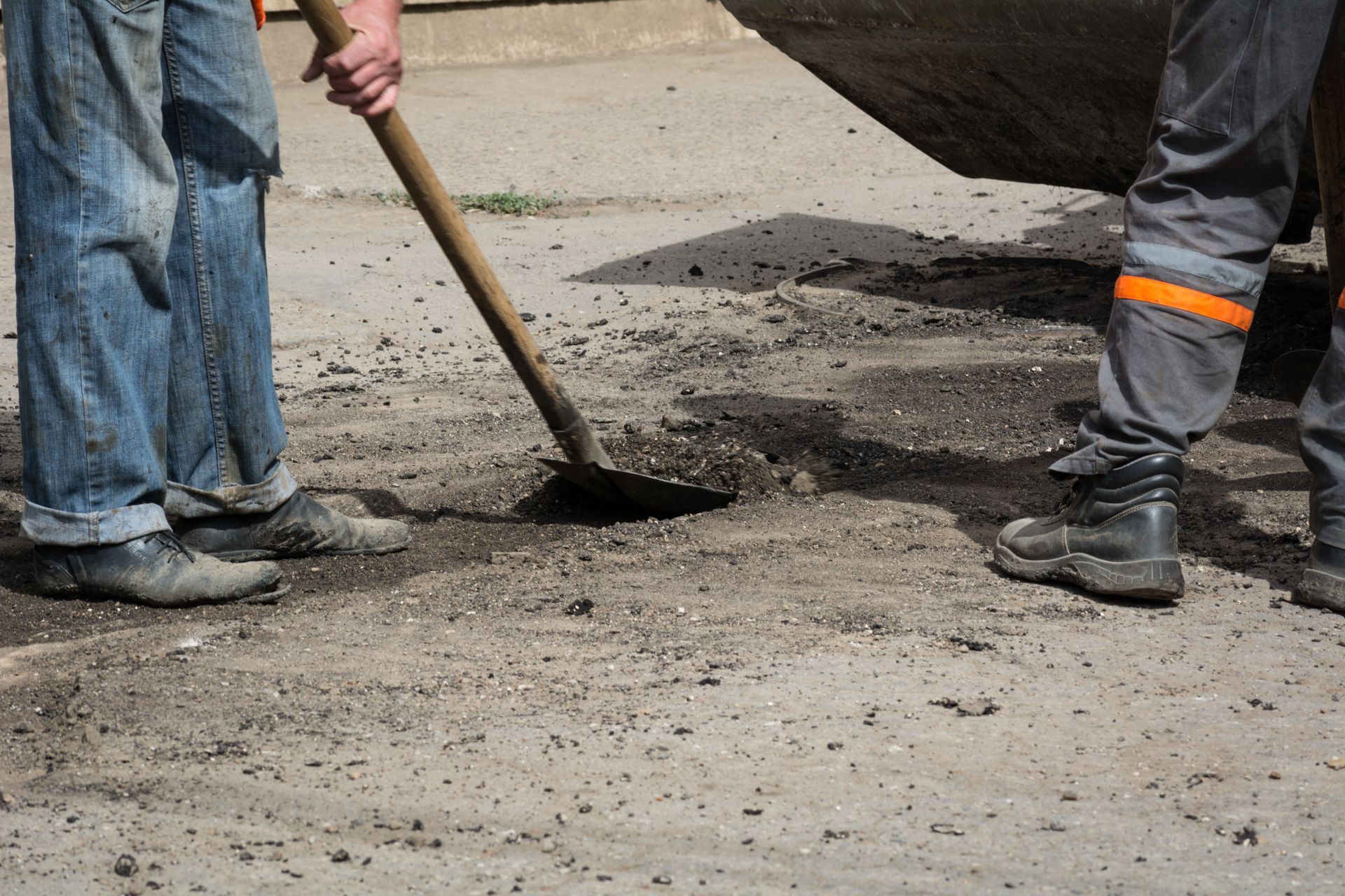 Workers patching a pothole with asphalt on a road, using a shovel near boots and a truck tire. Workers patching a pothole with asphalt on a road, using a shovel near boots and a truck tire.