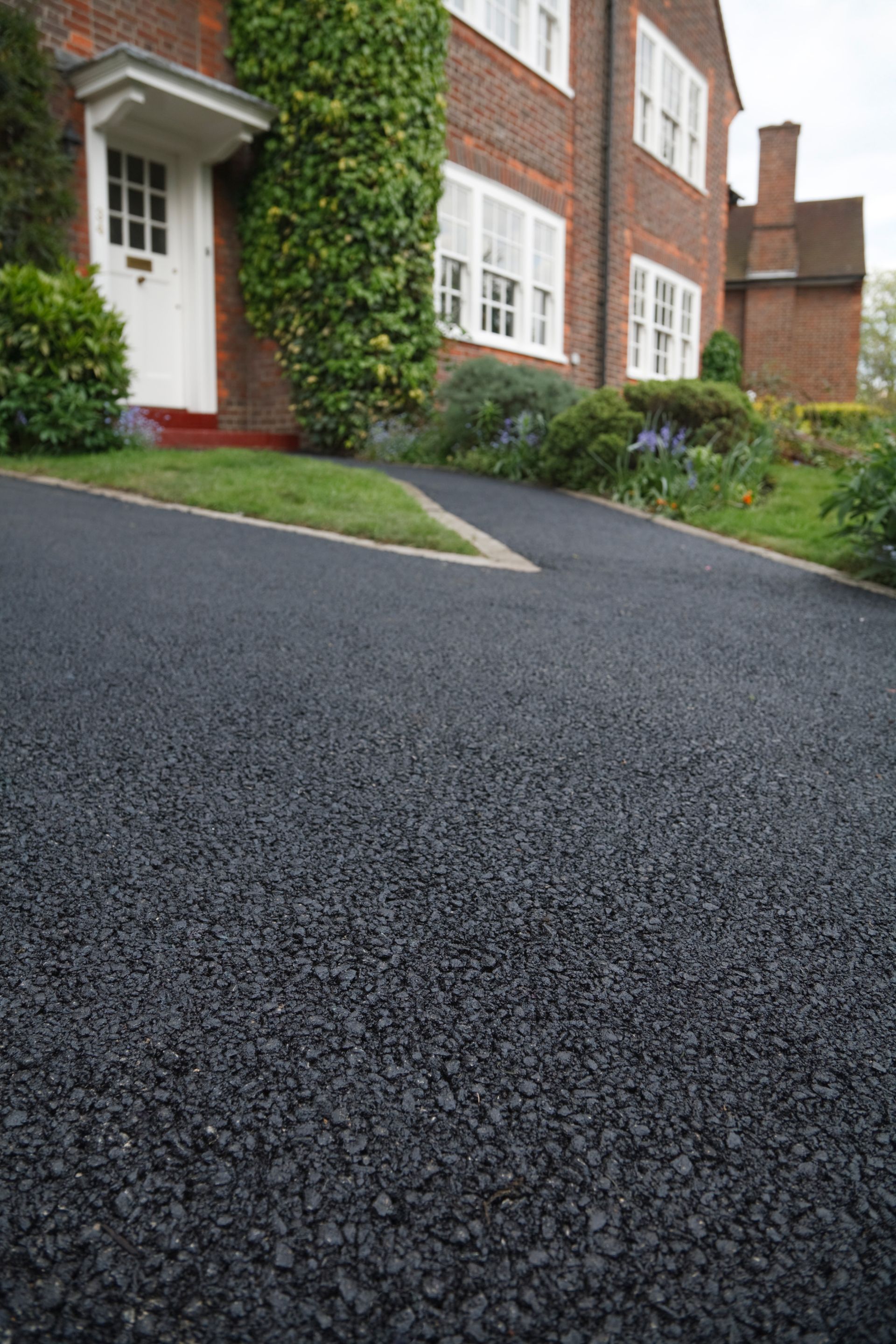 Low-angle view of a brick house with white trim and a sloped black driveway beside green shrubs. Low-angle view of a brick house with white trim and a sloped black driveway beside green shrubs.