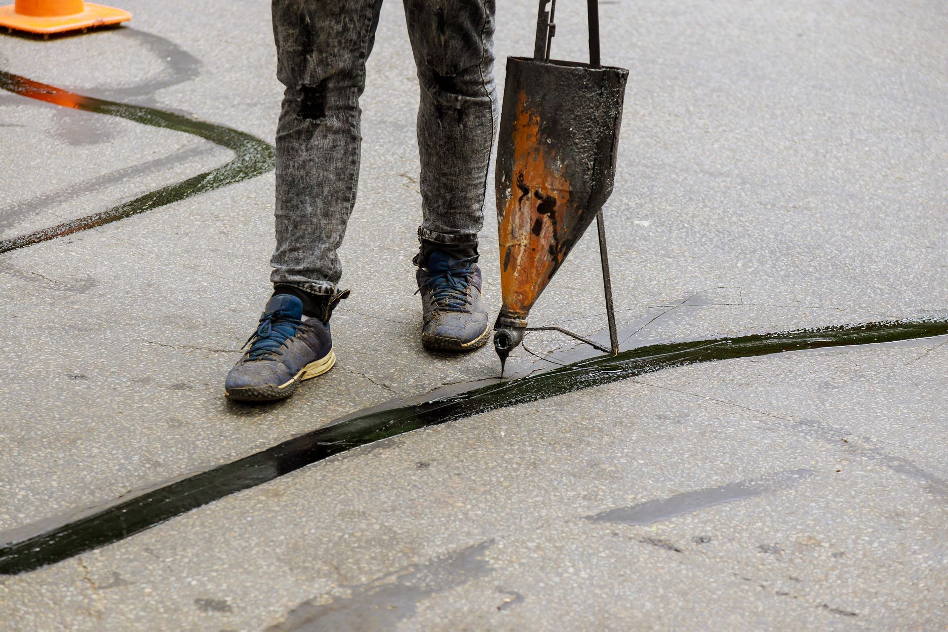 Person wearing jeans and sneakers holding a metal shovel on a wet pavement with crack repair sealant Person wearing jeans and sneakers holding a metal shovel on a wet pavement with crack repair sealant