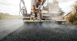 Road paving machine laying fresh black asphalt on a road, with dust and construction equipment visible Road paving machine laying fresh black asphalt on a road, with dust and construction equipment visible