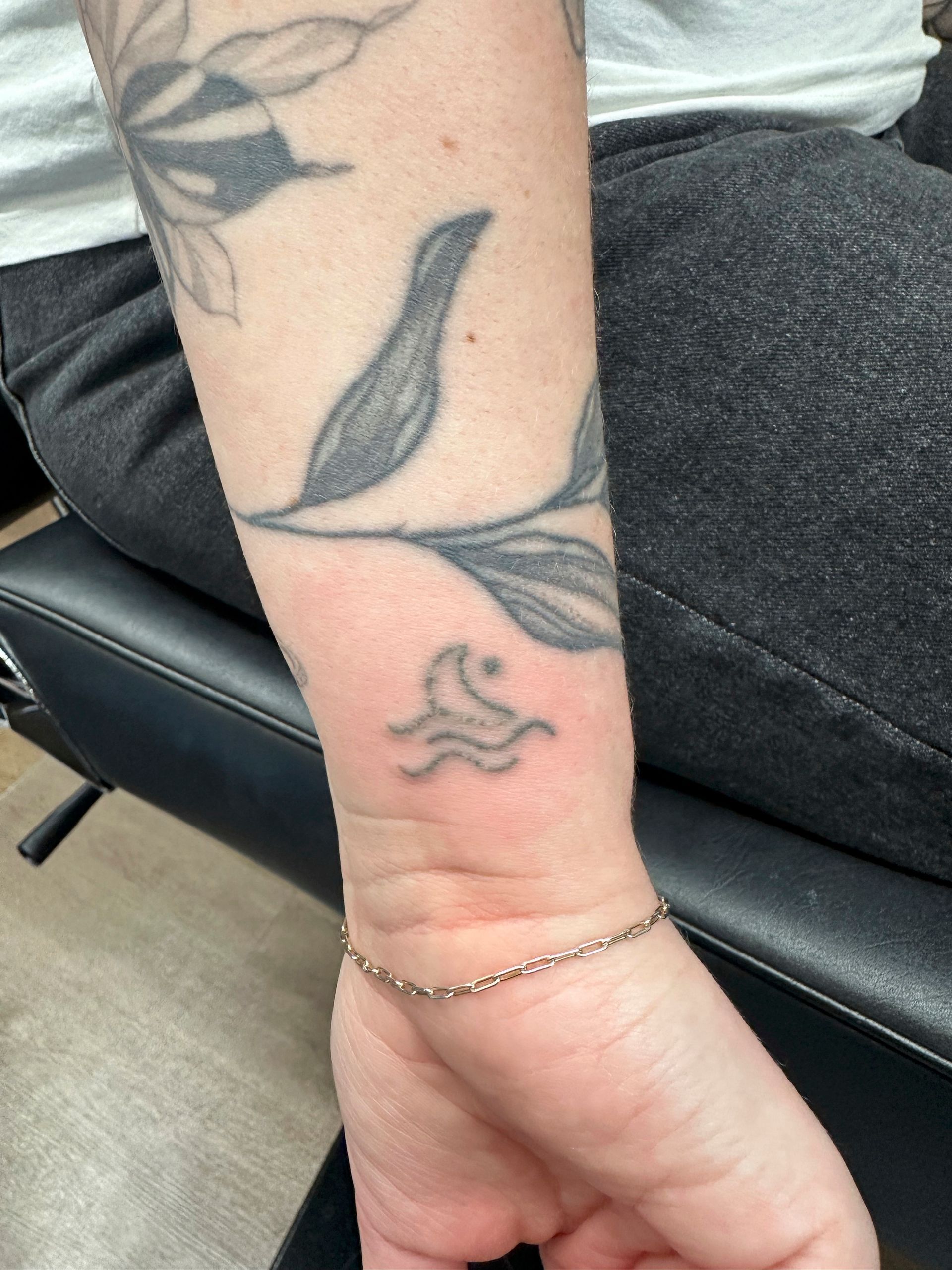 Een close-up van een pols met een kleine tatoeage van een halve maan boven golven, onder een grotere tatoeage van een blad.