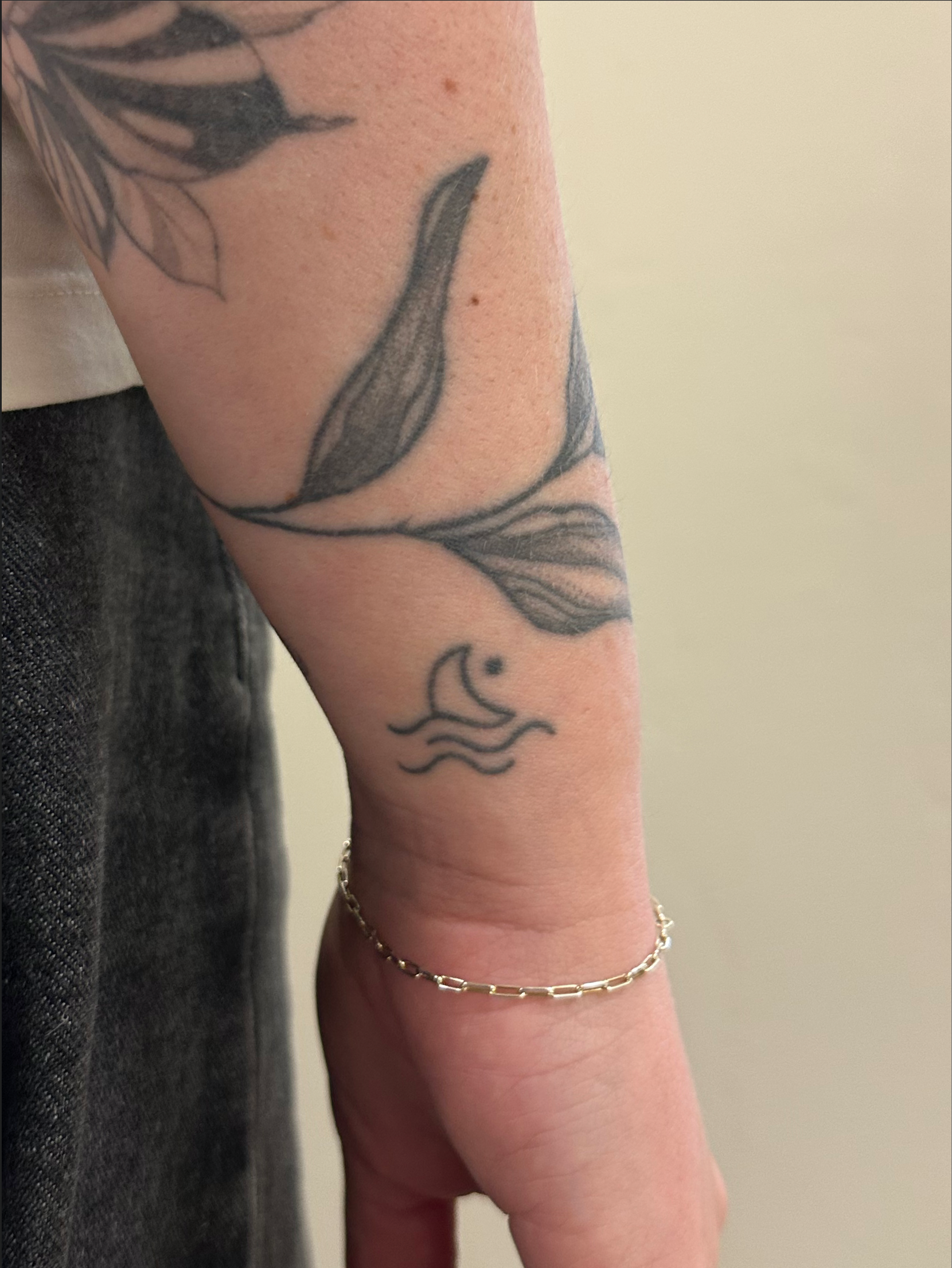 Een close-up van een onderarm met een zwarte inkt tatoeage van een halve maan boven golvende lijnen, met daarboven grotere bladmotieven.