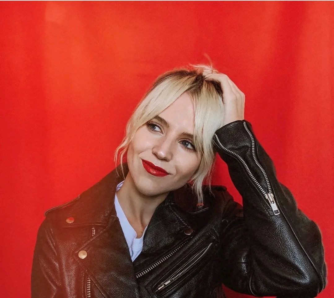 A person with blonde hair and red lipstick poses against a solid red background, wearing a black leather jacket.