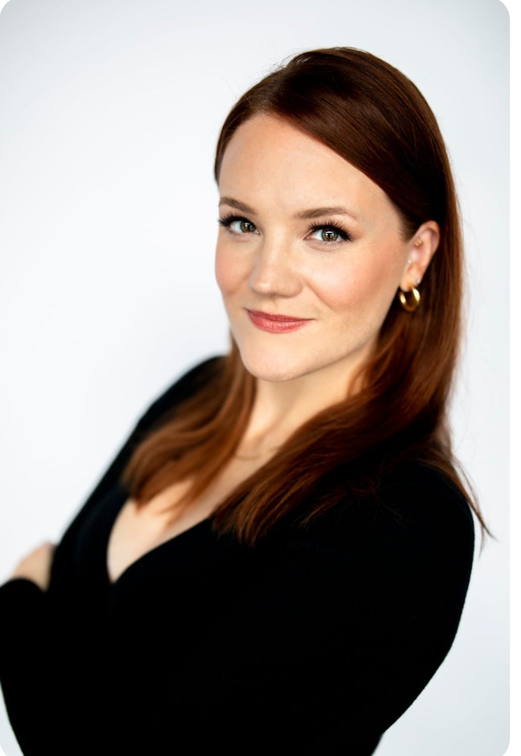 A person with shoulder-length auburn hair wears a black top, smiling slightly against a plain white background.