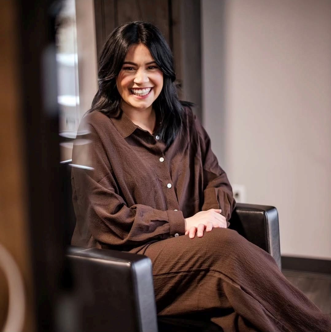 A smiling person with dark hair wears a brown button-up shirt and sits in a black chair indoors.