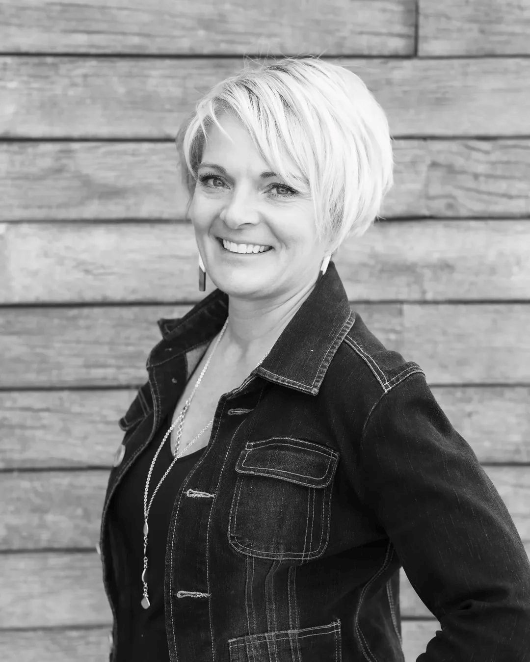 A person with a short, layered haircut smiles while posing in front of a wood-paneled wall, wearing a dark denim jacket.