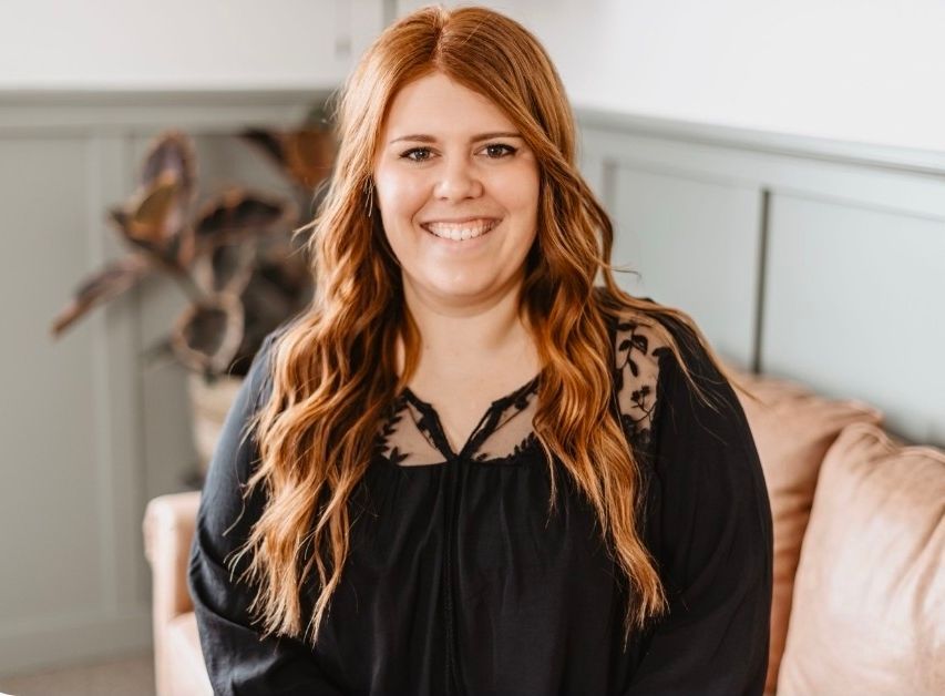 A smiling person with long auburn hair wearing a black blouse with lace detailing, seated in a room with paneled walls.