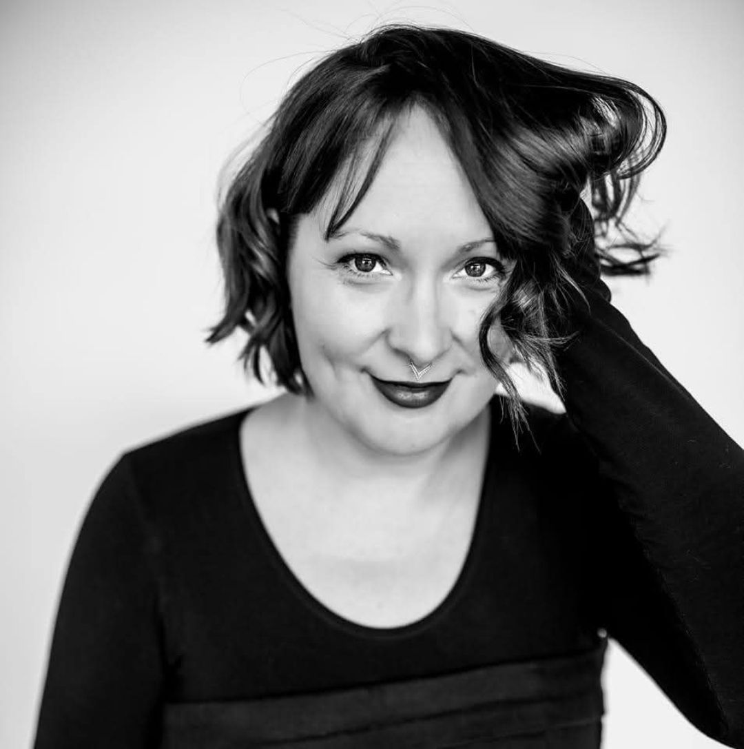 A person with dark hair and a septum piercing poses against a light background, smiling with one hand touching their hair.
