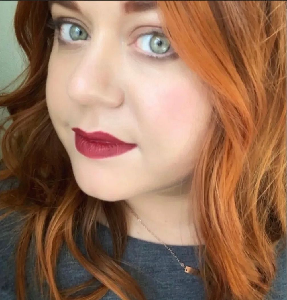 A close-up selfie of a person with wavy, ginger hair, bright green eyes, and dark berry-colored lipstick.