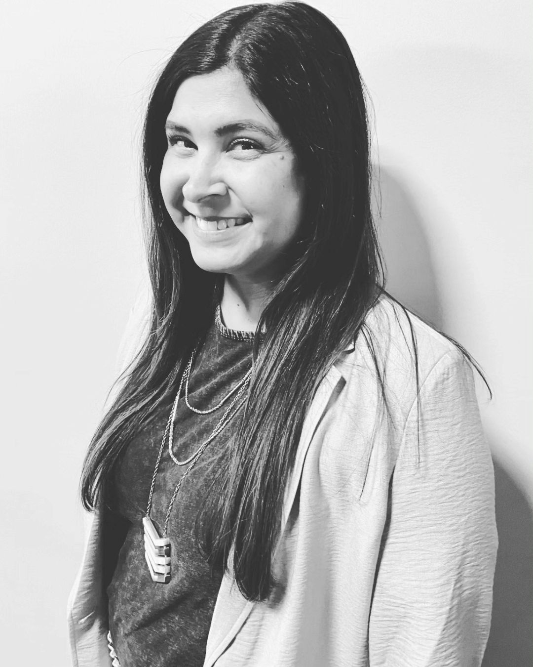 A smiling person with long dark hair, wearing a necklace and a blazer, standing against a plain light-colored wall.
