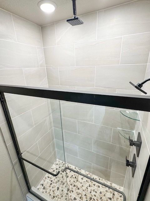 Shower with glass door and black fixtures; light beige tiles, pebble floor.