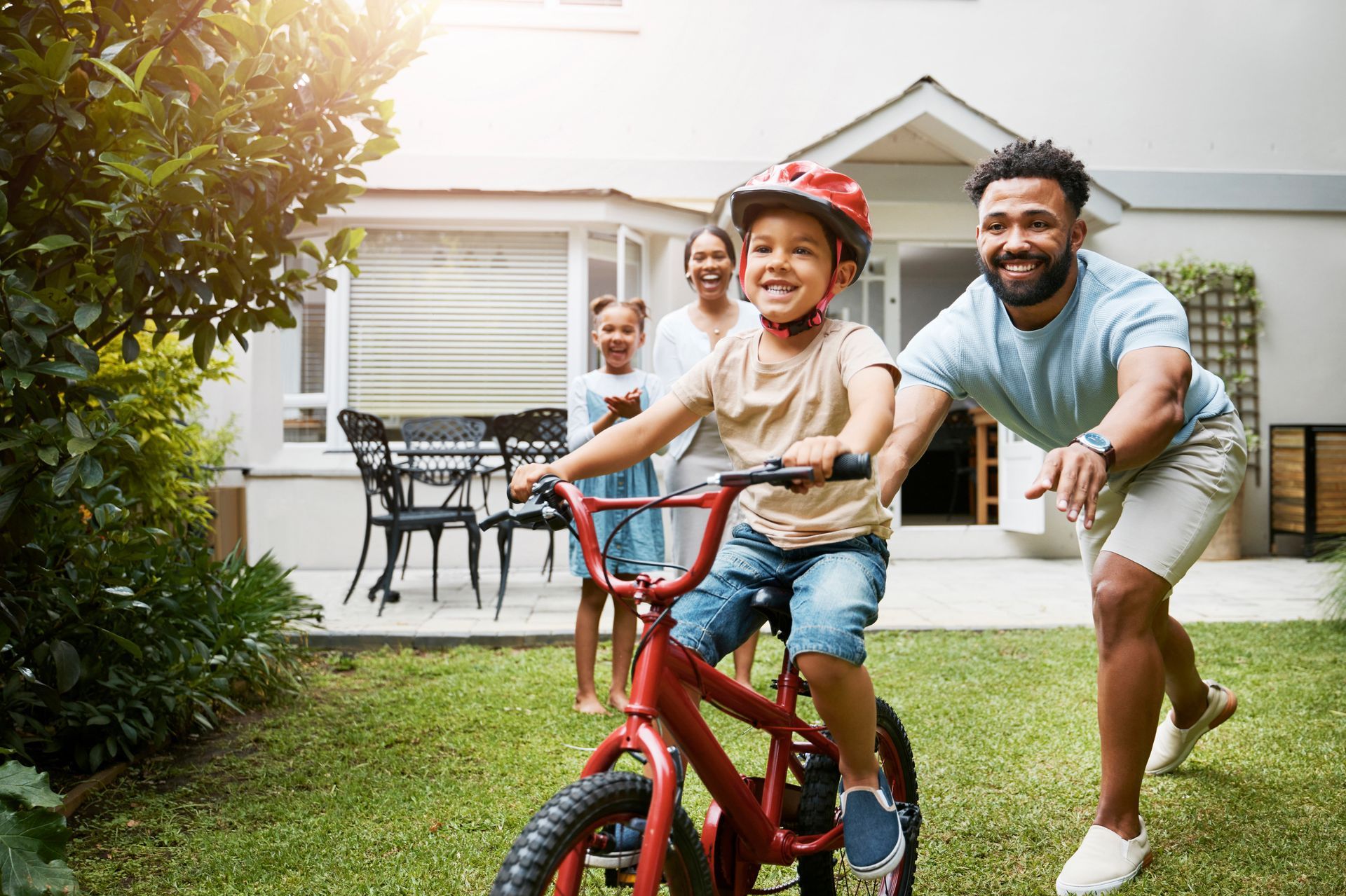 Boy rides a red bike in backyard, father runs alongside. Family watches.