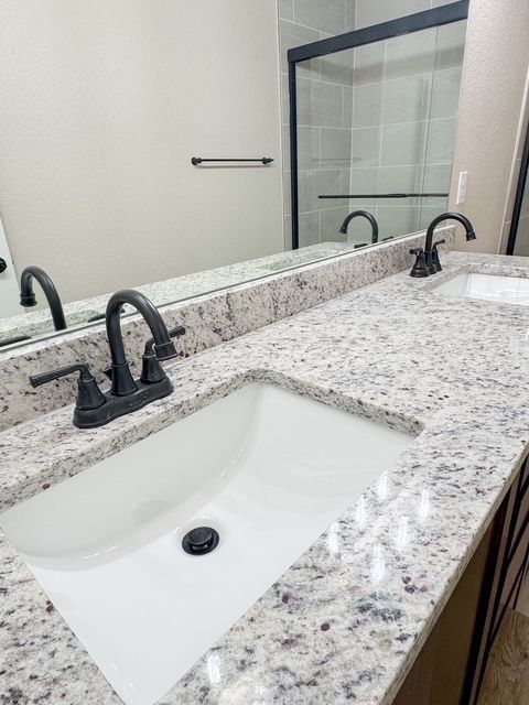 Bathroom with double sinks, granite countertop, black faucets, large mirror, and a glass shower door.