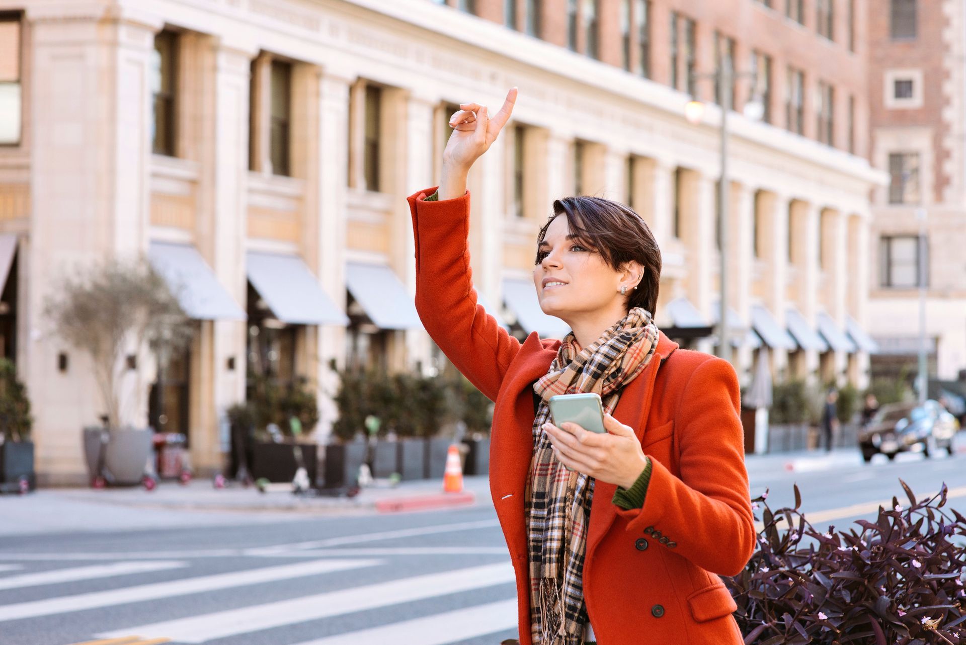 Woman in orange coat hailing a cab on a city street, holding phone, looking up.