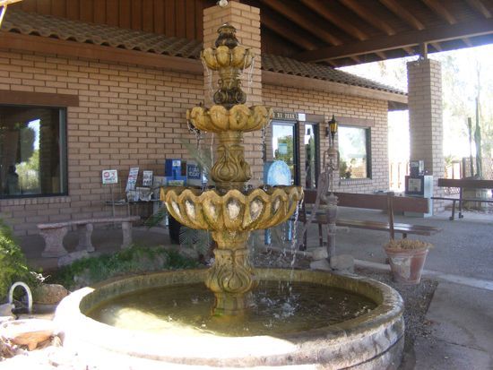 Ornate tiered fountain in front of a brick building with a covered walkway and benches.
