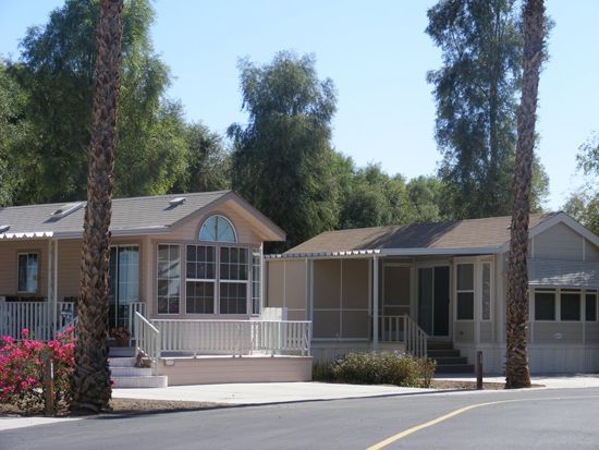 Two beige homes with porches, palm trees in the foreground, and lush greenery in the background on a sunny day.