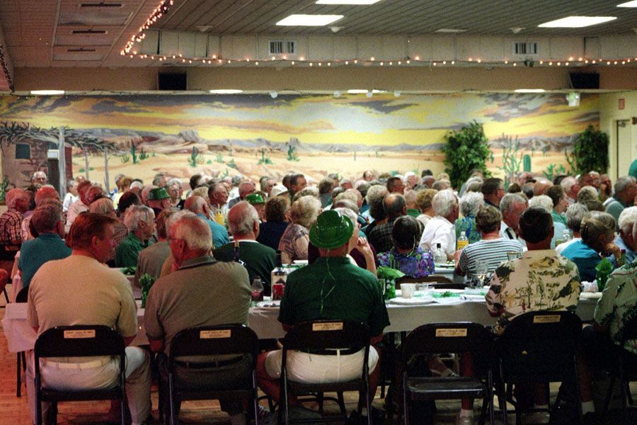 Large group of people seated at tables in a banquet hall, some wearing green, with a desert mural on the back wall.
