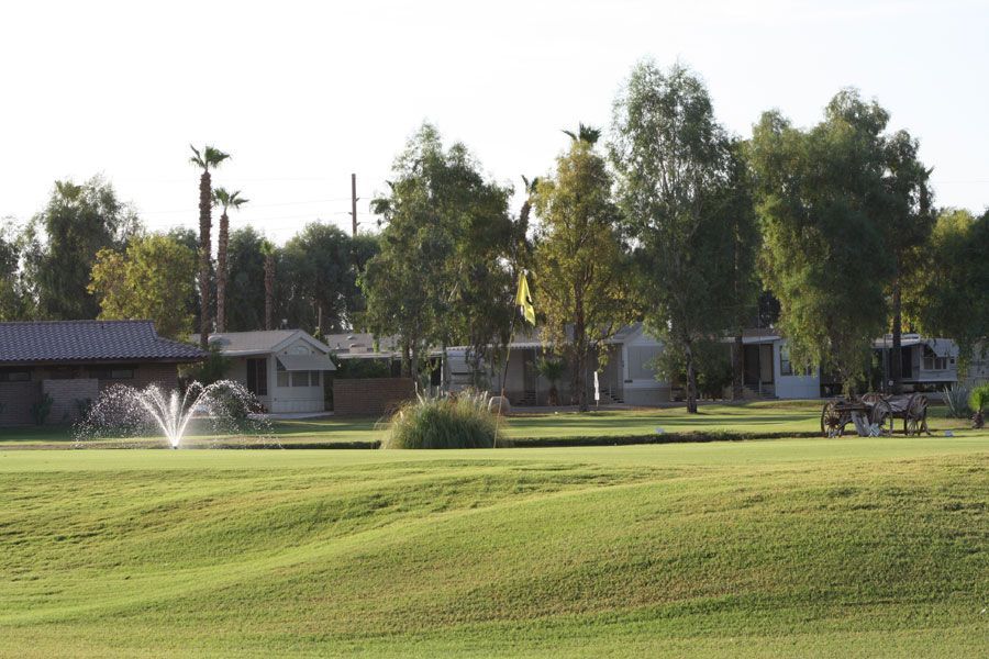 Green golf course with a fountain and homes in the background under a sunny sky.