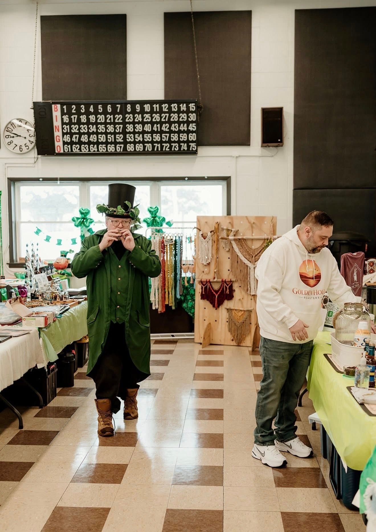 A person in a leprechaun costume and another in a hoodie stand in a hall with a bingo board and craft displays.