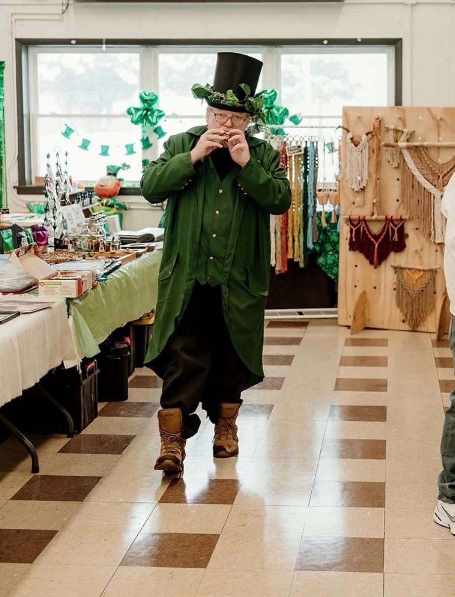 A person wearing a green coat and a top hat with clover decorations walks through a room, holding an item to their mouth.