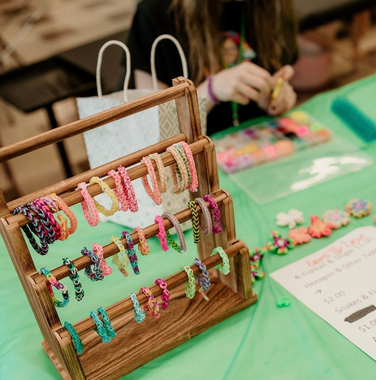 A wooden stand displaying colorful handmade rubber band bracelets on a green table, with a person crafting in the back.