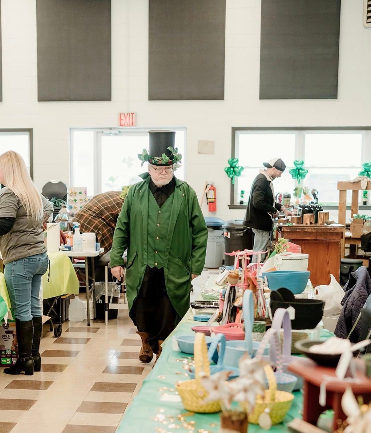 A person dressed as a leprechaun in a top hat and green coat walks through a room filled with tables of items for sale.