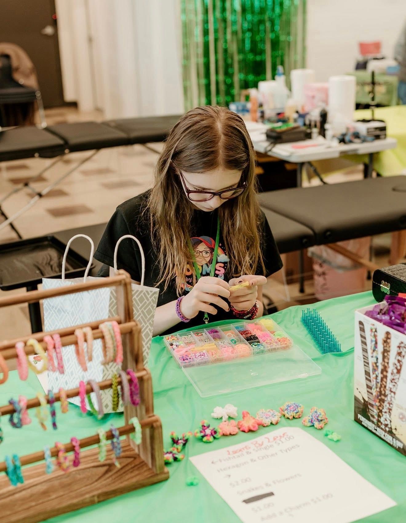 A person works on a craft project at a table with a display rack of colorful loom-band bracelets and a price list.