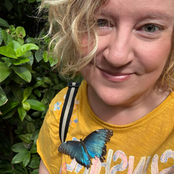 A person with shoulder-length blonde hair wears a plaid scarf and red lipstick in front of a decorated Christmas tree.