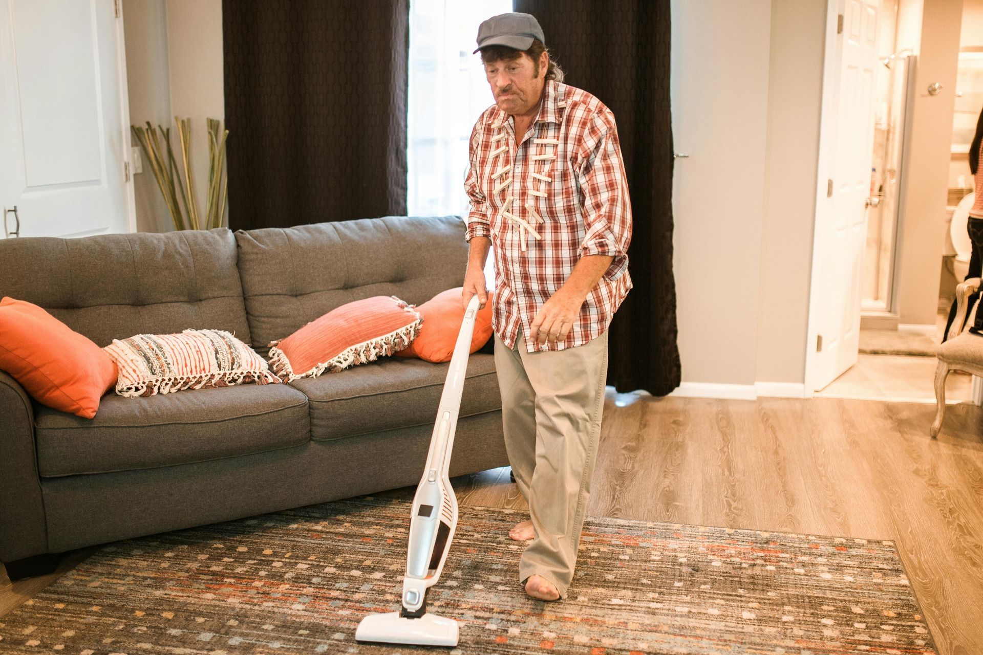 A man in a plaid shirt and cap uses a stick vacuum on a rug in a living room.