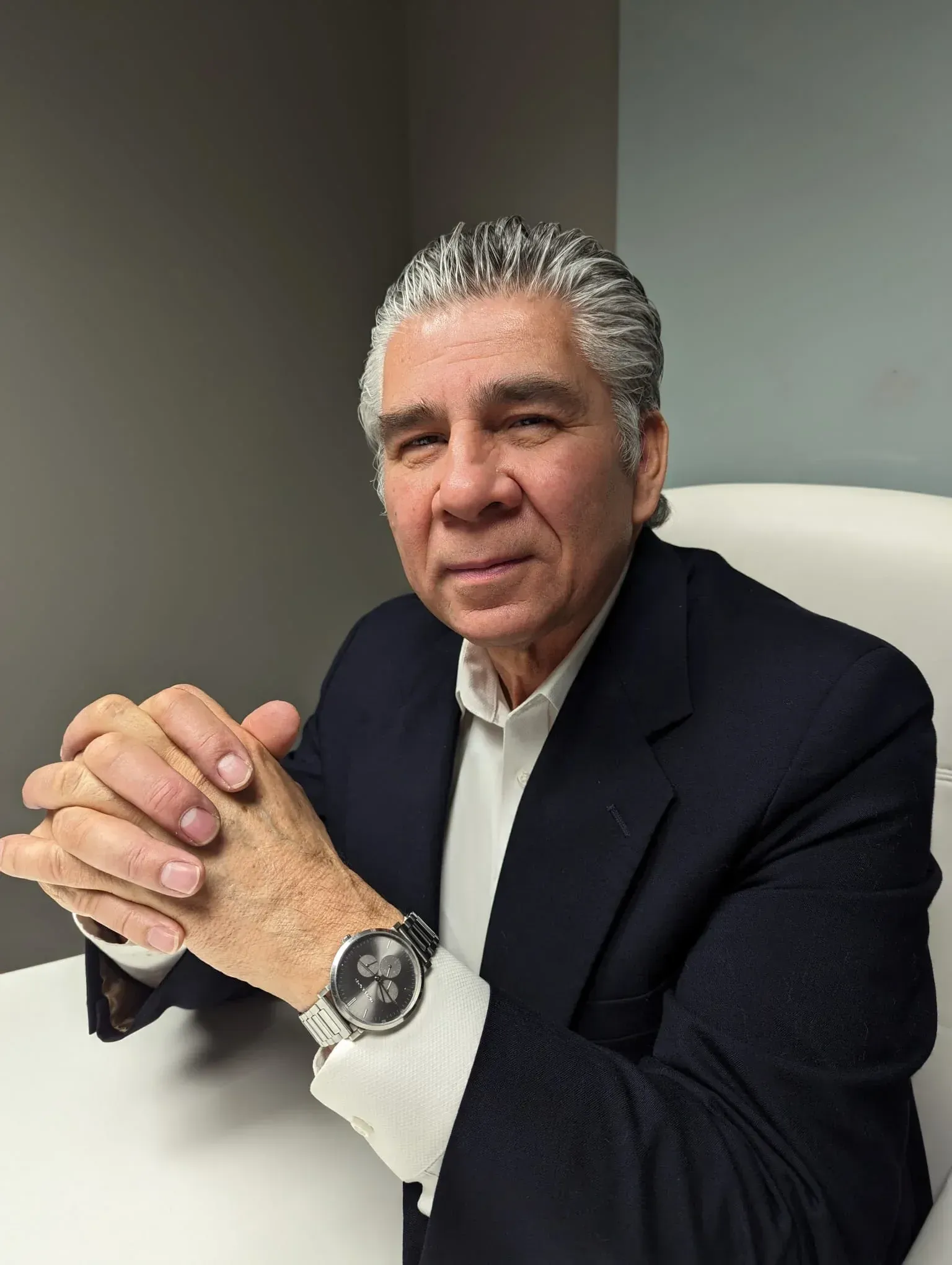 Man in a dark blazer seated at a desk, hands clasped, wearing a wristwatch, in an office setting.
