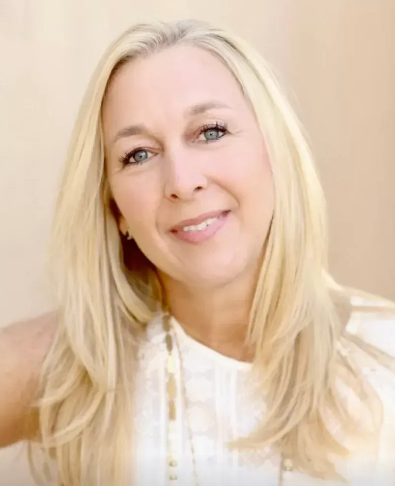 A woman with long blonde hair is wearing a white shirt and smiling.