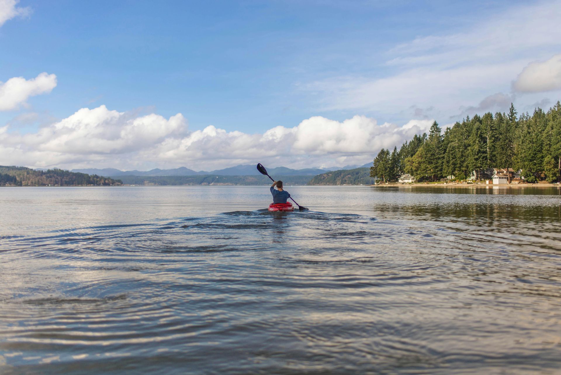 Kayaker in a red jacket paddling on a calm lake with forested shoreline and mountains in the distance