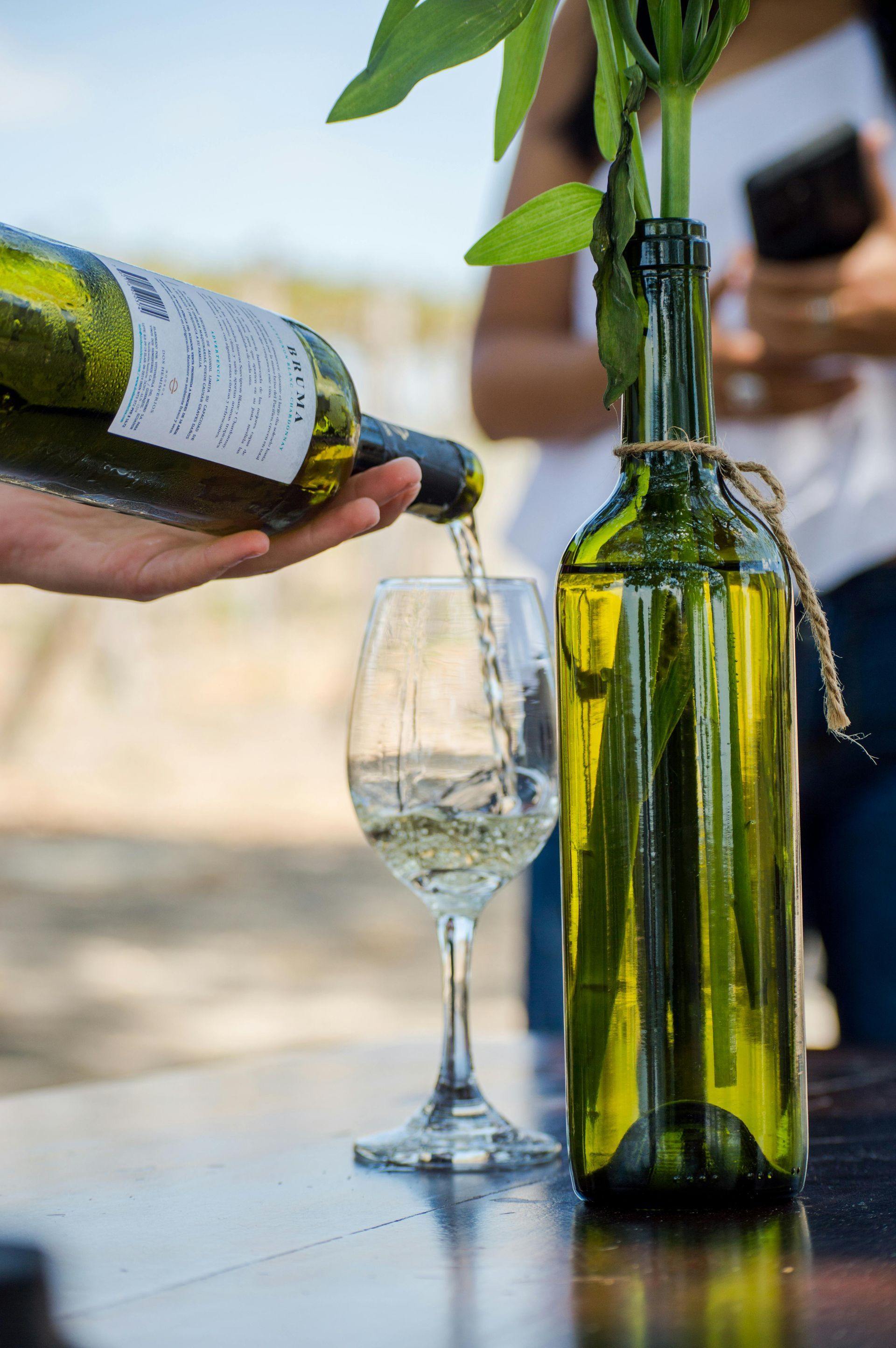 Wine being poured into a glass beside a green bottle on a table outdoors