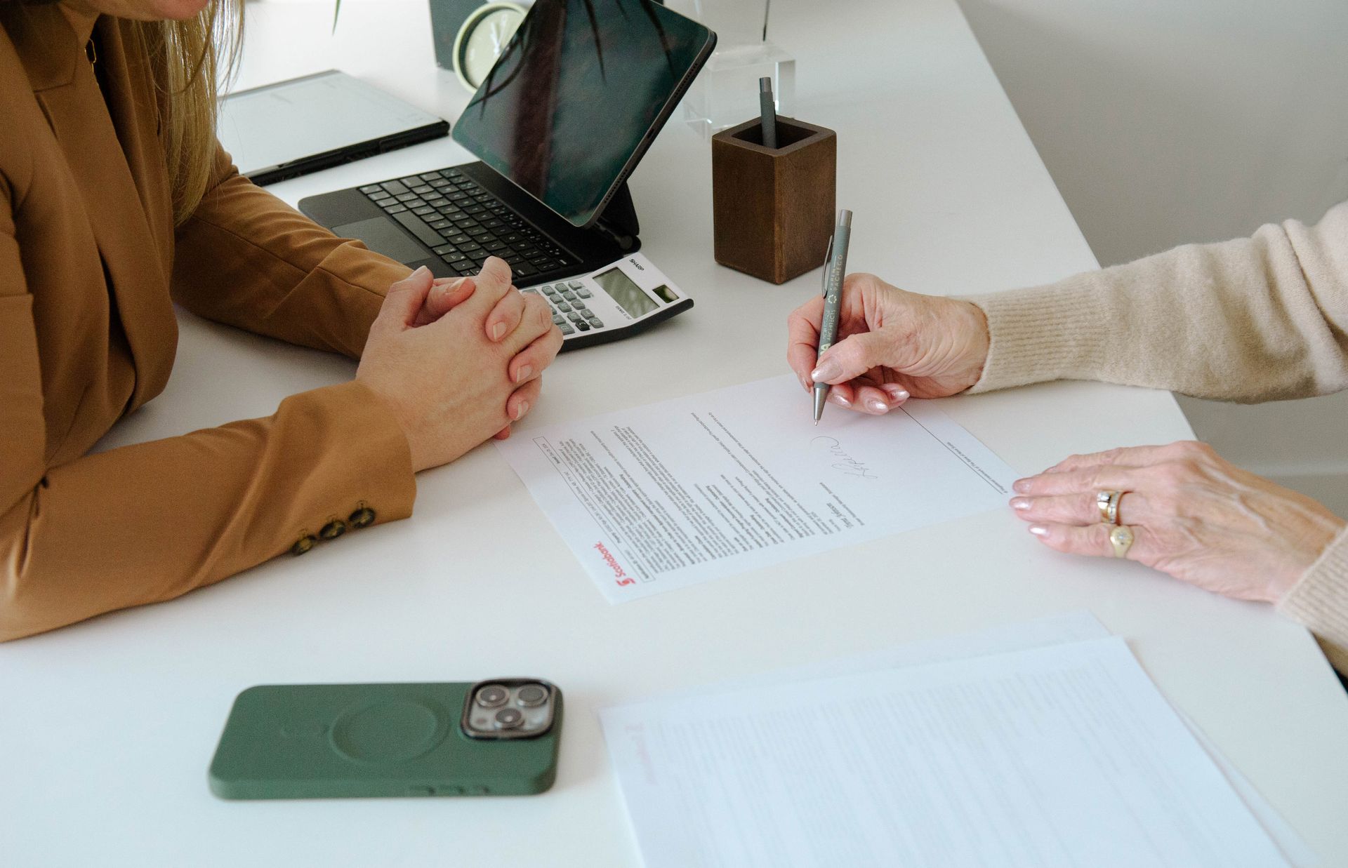 Two people at a desk; one signs a document while the other watches.