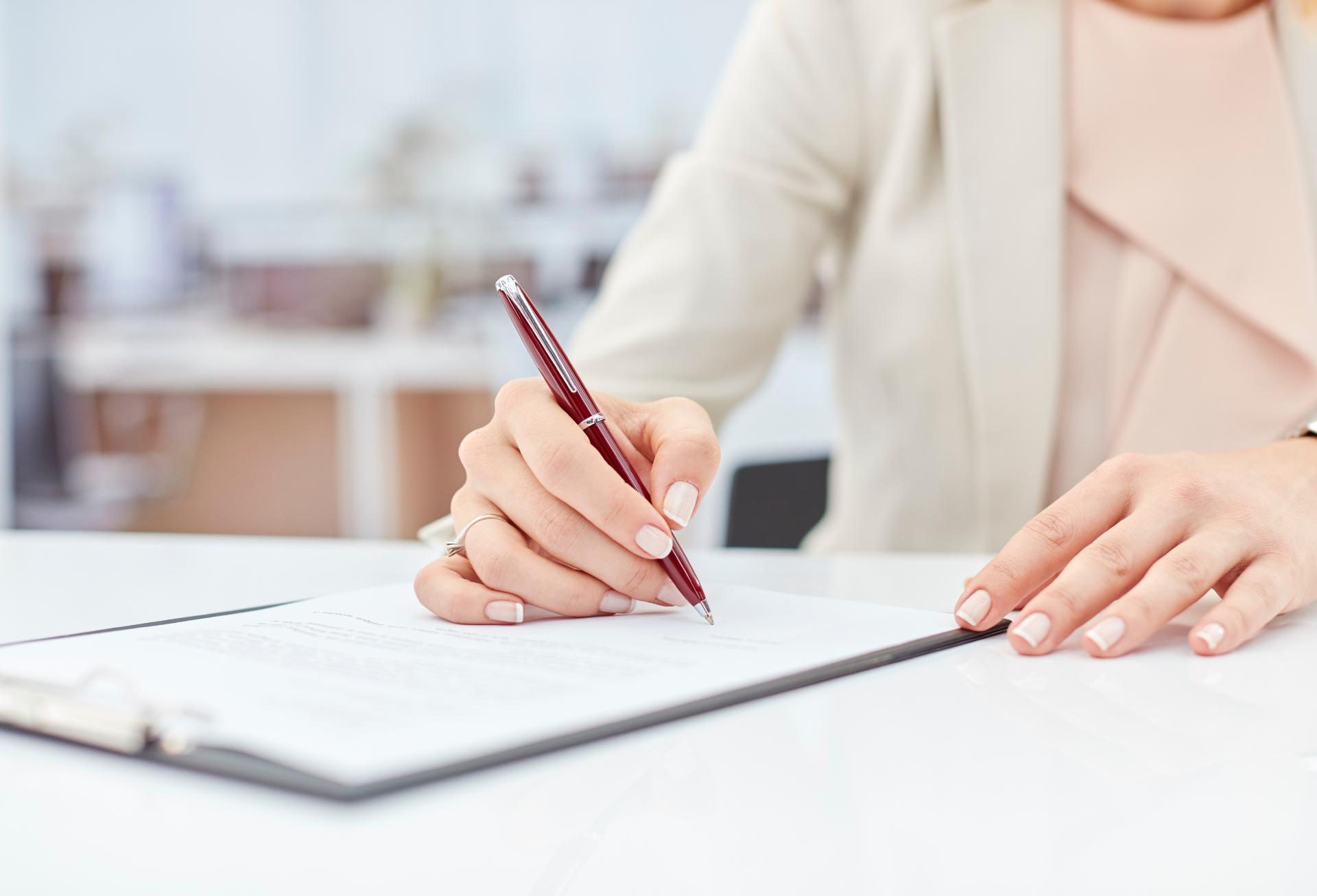 Woman's hand writing with a pen on paper in a clipboard, wearing a beige blazer.
