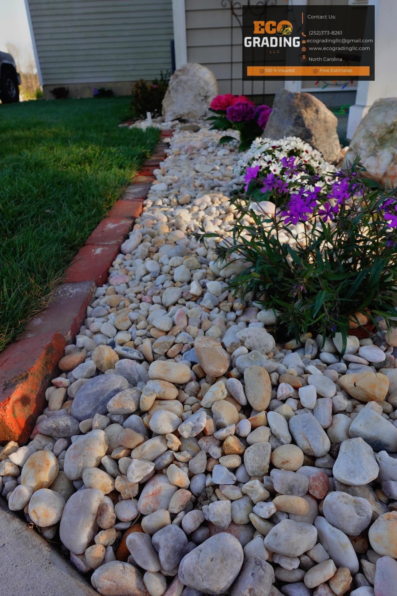 Landscaped flower bed with tan rocks and flowers, edged by bricks, beside a lawn.