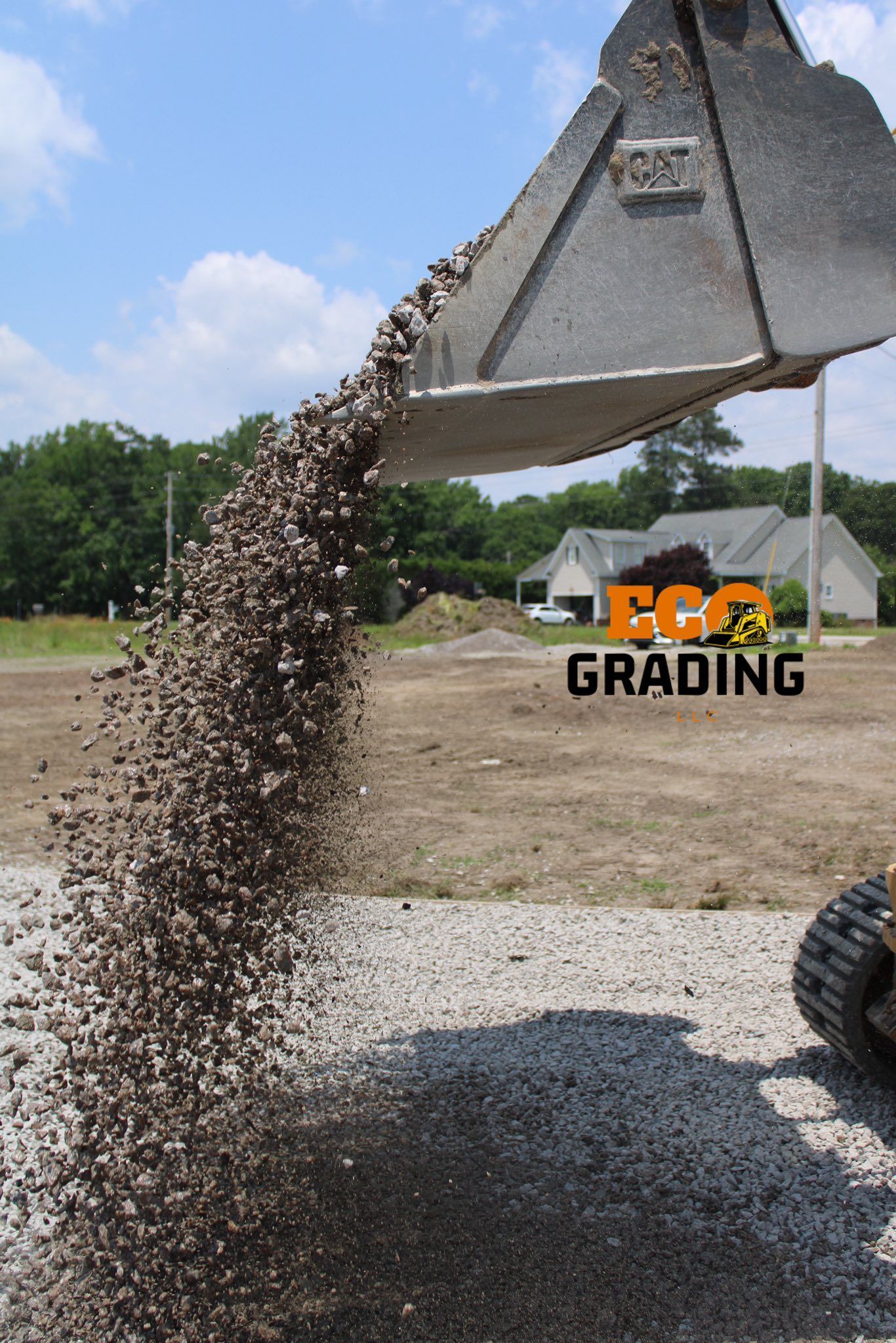 Excavator dumping gravel onto a dirt surface under a blue sky, near a residential area.