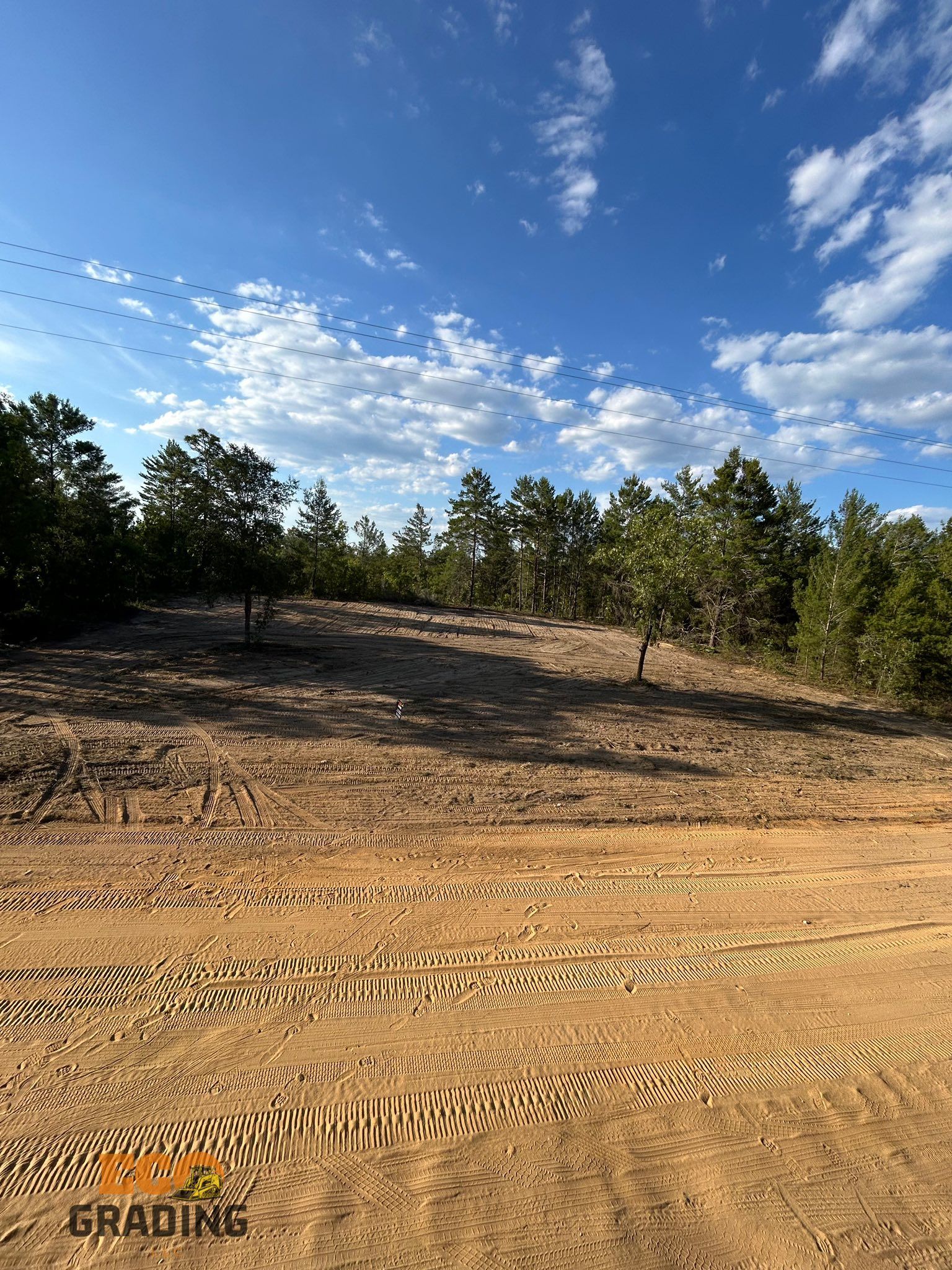 A cleared dirt lot with tire tracks, trees in the background, and a cloudy blue sky above.