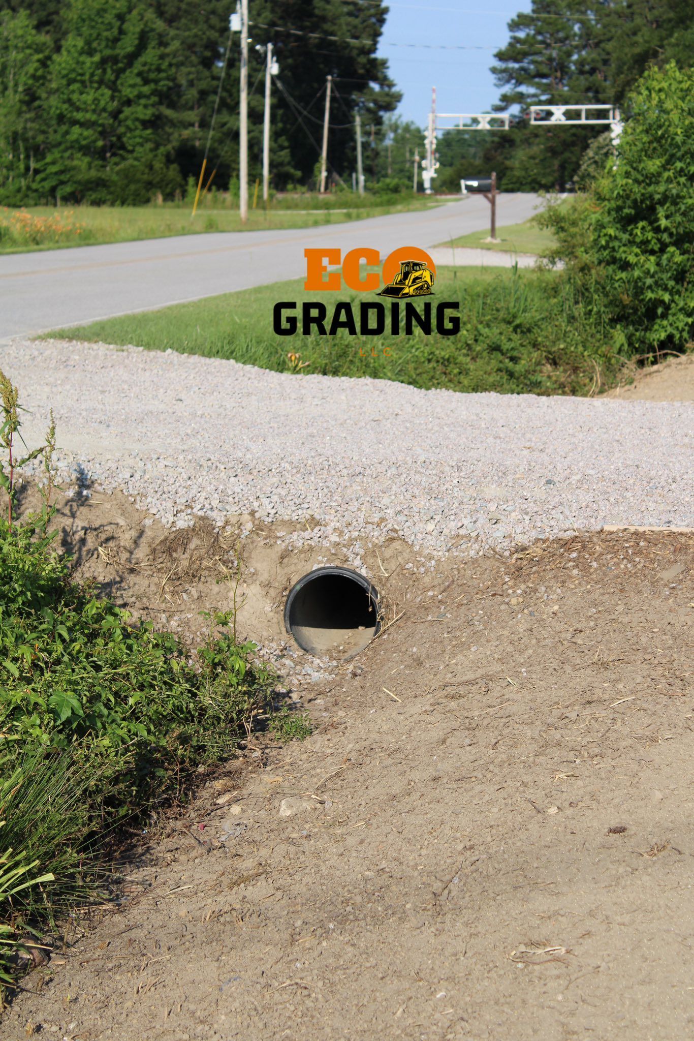 A culvert under a gravel road, with vegetation on either side. A road and trees in the background.