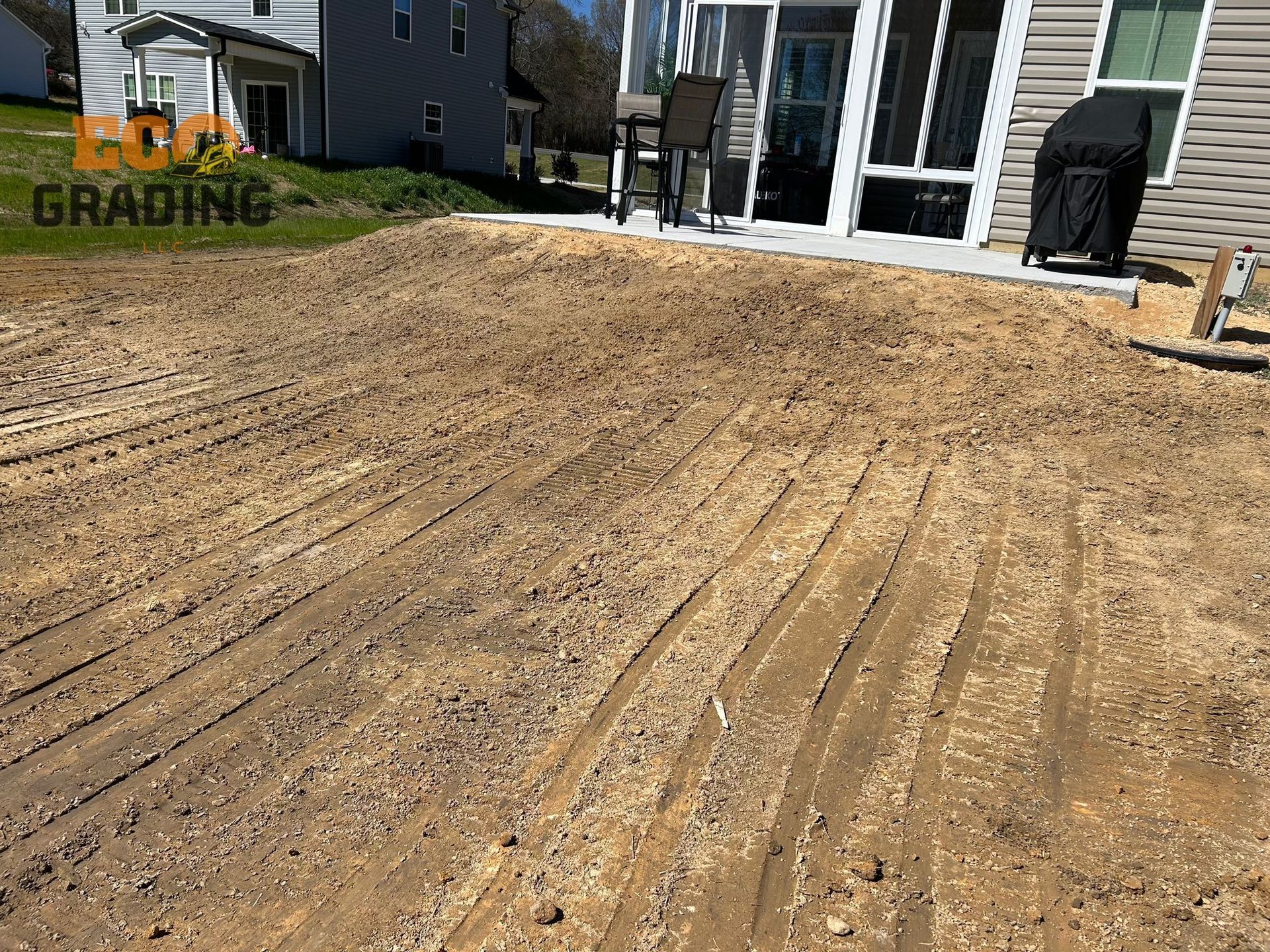 Graded yard with gravel, next to a patio and house. Earth-toned colors. Tracks from equipment visible.