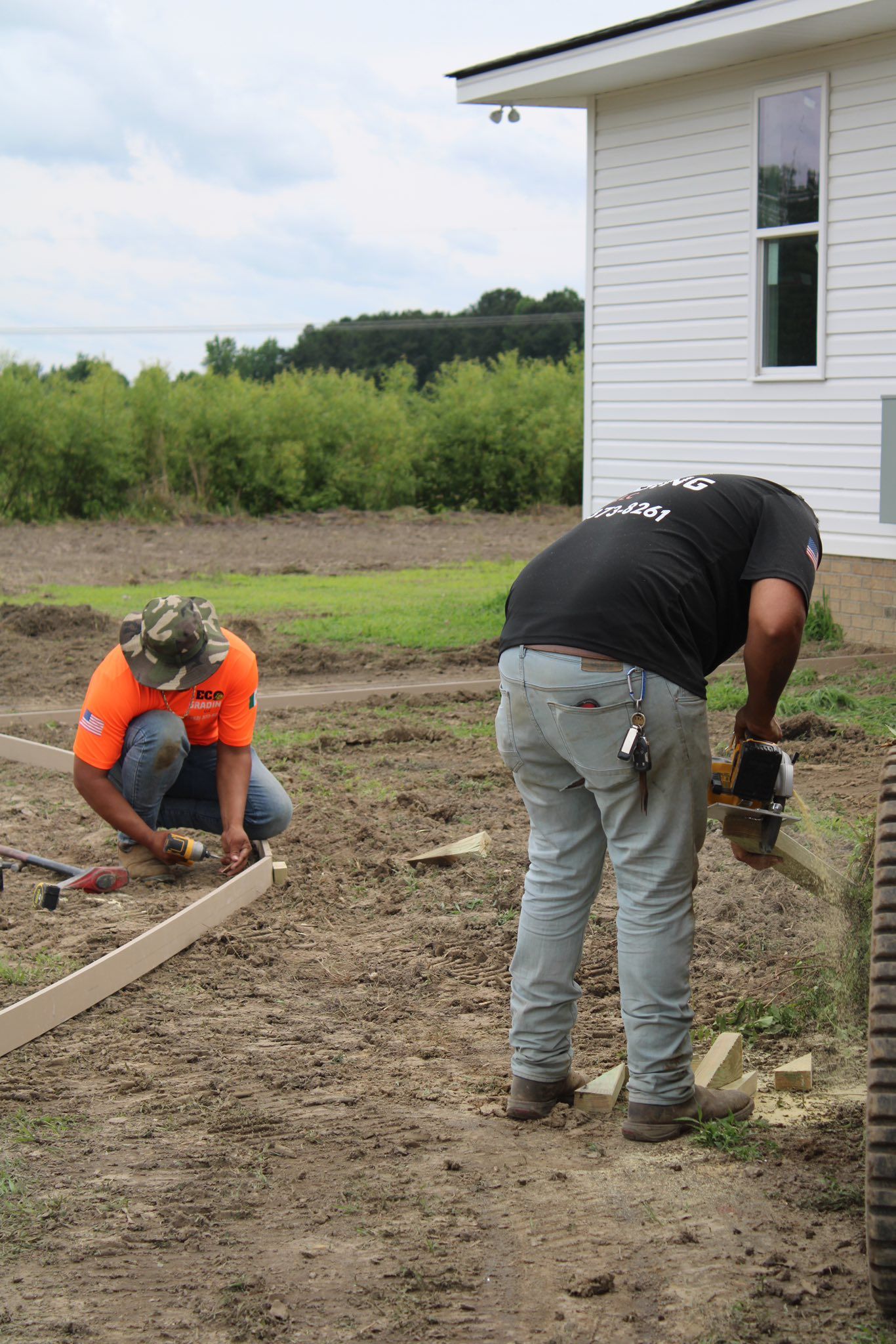 Two construction workers installing a border next to a white building; one cuts, one works on the ground.